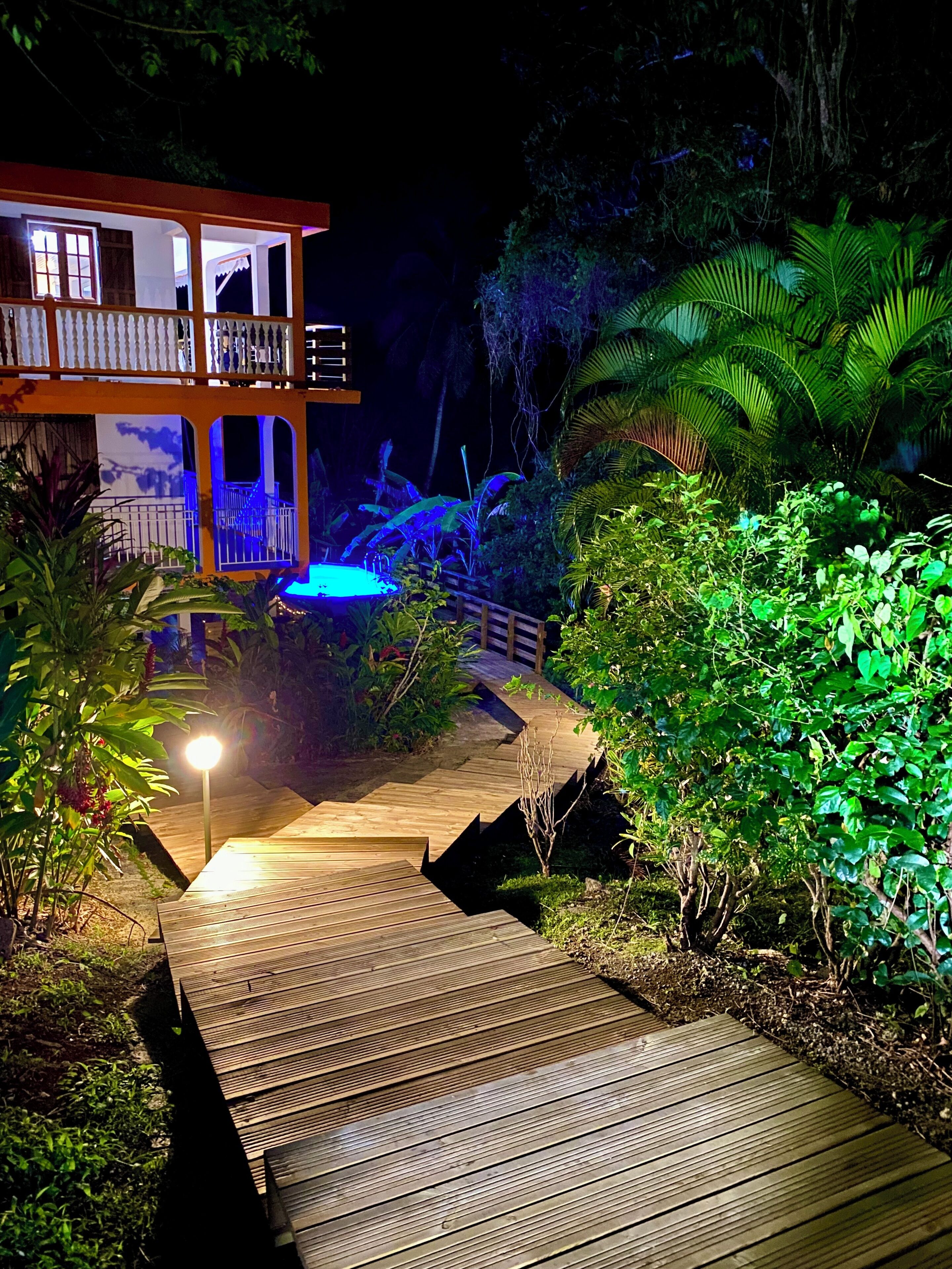 Una terraza de madera que conduce a un edificio de dos pisos con balcones, rodeado de abundante vegetación e iluminado con luces azules.
