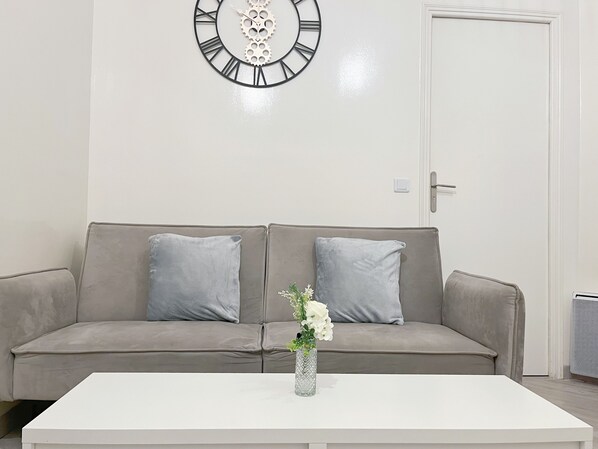 Een moderne woonkamer met een beige bank, twee grijze kussens, een witte salontafel en een decoratieve wandklok.
