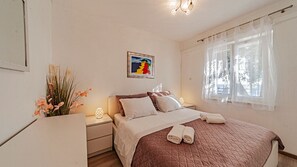 Ein Schlafzimmer mit Bett, Nachttischen, einem Fenster mit durchscheinenden Vorhängen und einem Wandbild.