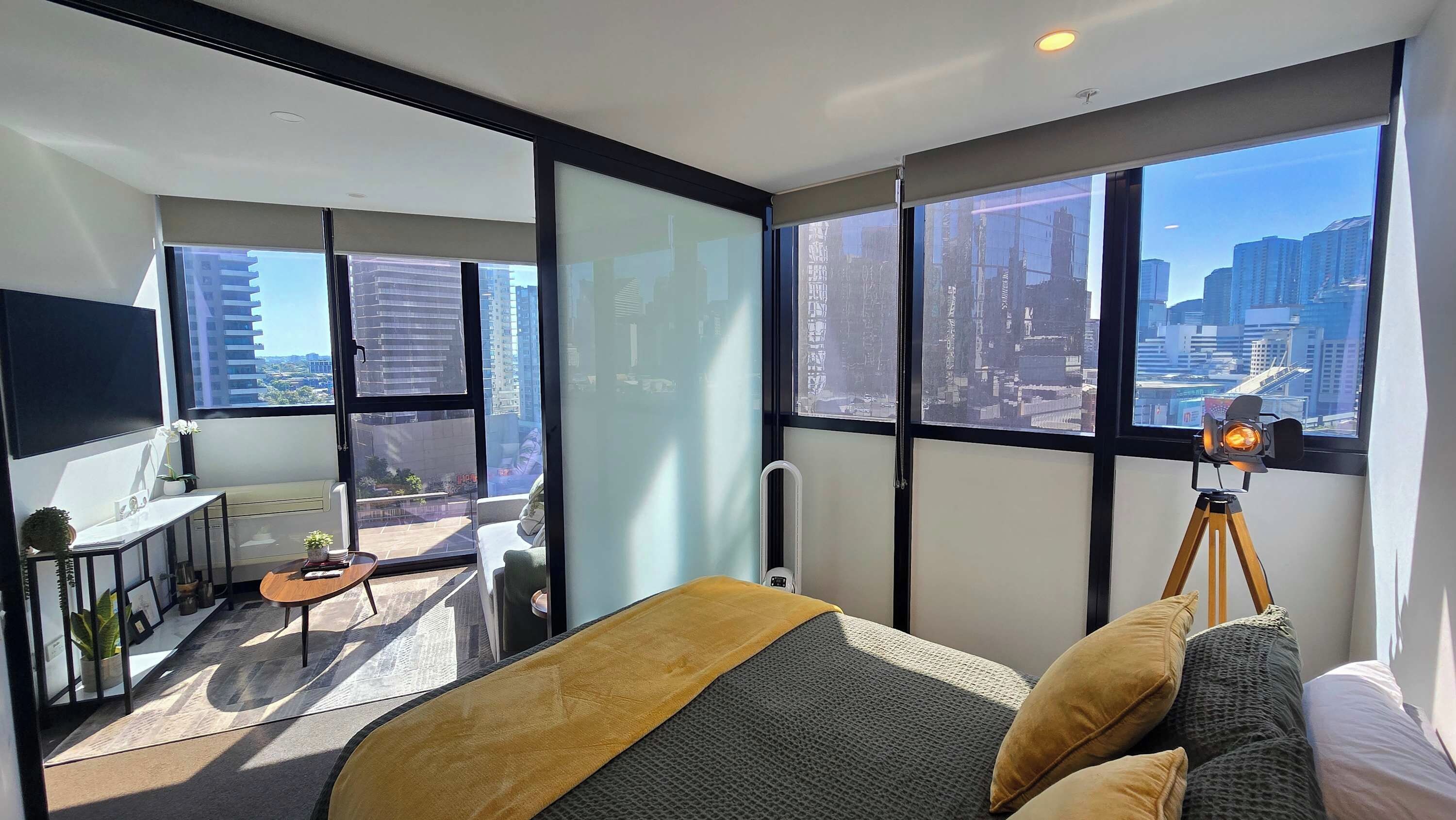 A modern hotel room with a large bed, a small desk, a chair, a balcony with a view of the city, and a camera on a tripod.
