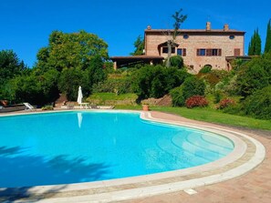 Uma piscina grande e sinuosa com borda de pedra, rodeada por um jardim bem cuidado e um edifício de pedra com telhado em azulejos.