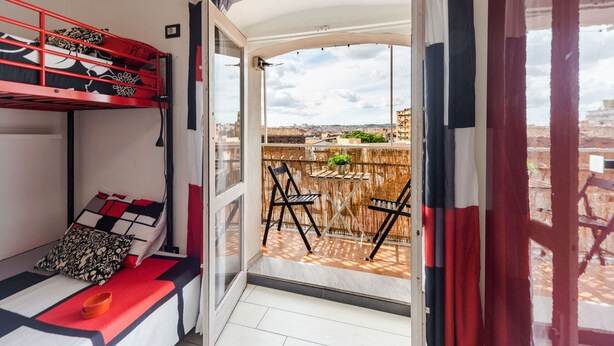 Una habitación con literas, una pequeña mesa con sillas y vistas a la ciudad desde un balcón abierto.