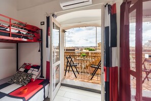 Quarto com beliche, uma pequena mesa com cadeiras e vista para a cidade através de uma varanda aberta.
