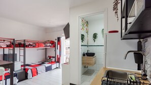 Una habitación pequeña con literas, un rincón cocina con pileta y encimera de madera, y vista al exterior a través de un ventanal.