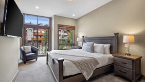 Una camera da letto con un letto grande, una sedia, un comodino con una lampada, una TV montata a parete e una finestra con vista sugli edifici.