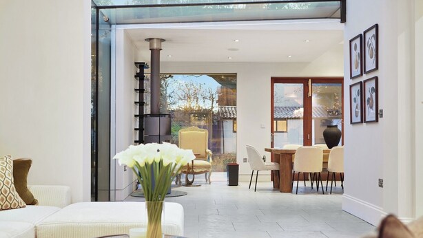 A modern living room with a glass wall, a fireplace, a dining area with a table and chairs, and a vase with flowers on a coffee table. - GRAND MANOR HOUSE WIMBLEDON (London)