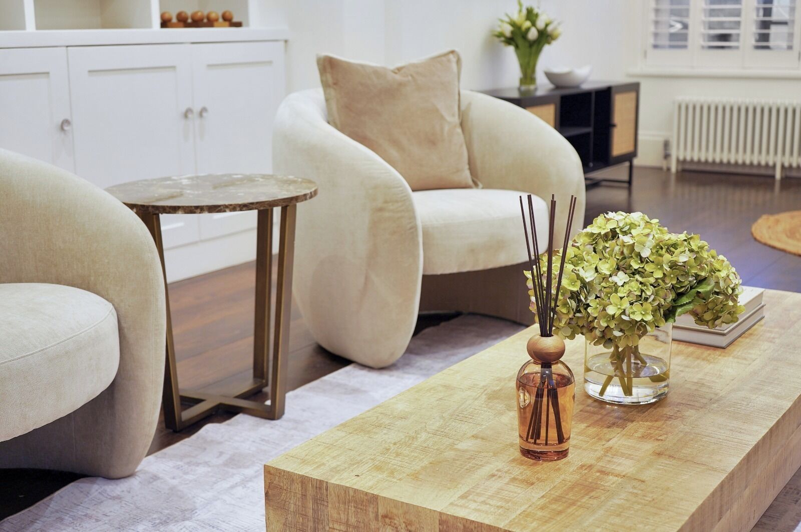 Een woonkamer met een beige bank, een klein rond tafeltje en een houten salontafel met twee vazen vol bloemen.
