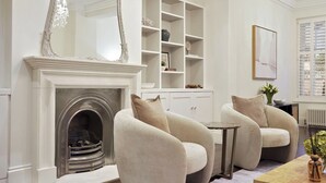 A living room with a fireplace, two armchairs, a coffee table, a bookshelf, and a chandelier. - GRAND MANOR HOUSE WIMBLEDON (London)