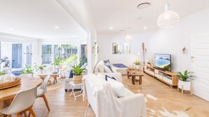 Een ruime woonkamer met een eettafel, banken en een televisie. Er staan planten en een groot raam zorgt voor natuurlijk licht.