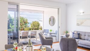 Un salon moderne avec un canapé gris, un coin repas avec une table dressée pour deux personnes, et un balcon offrant une vue sur la mer et la verdure.