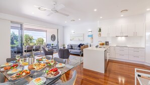 Une cuisine moderne avec des meubles blancs et un îlot central, un coin repas avec une table et des chaises en verre, et une vue sur l’extérieur grâce à de grandes portes coulissantes.