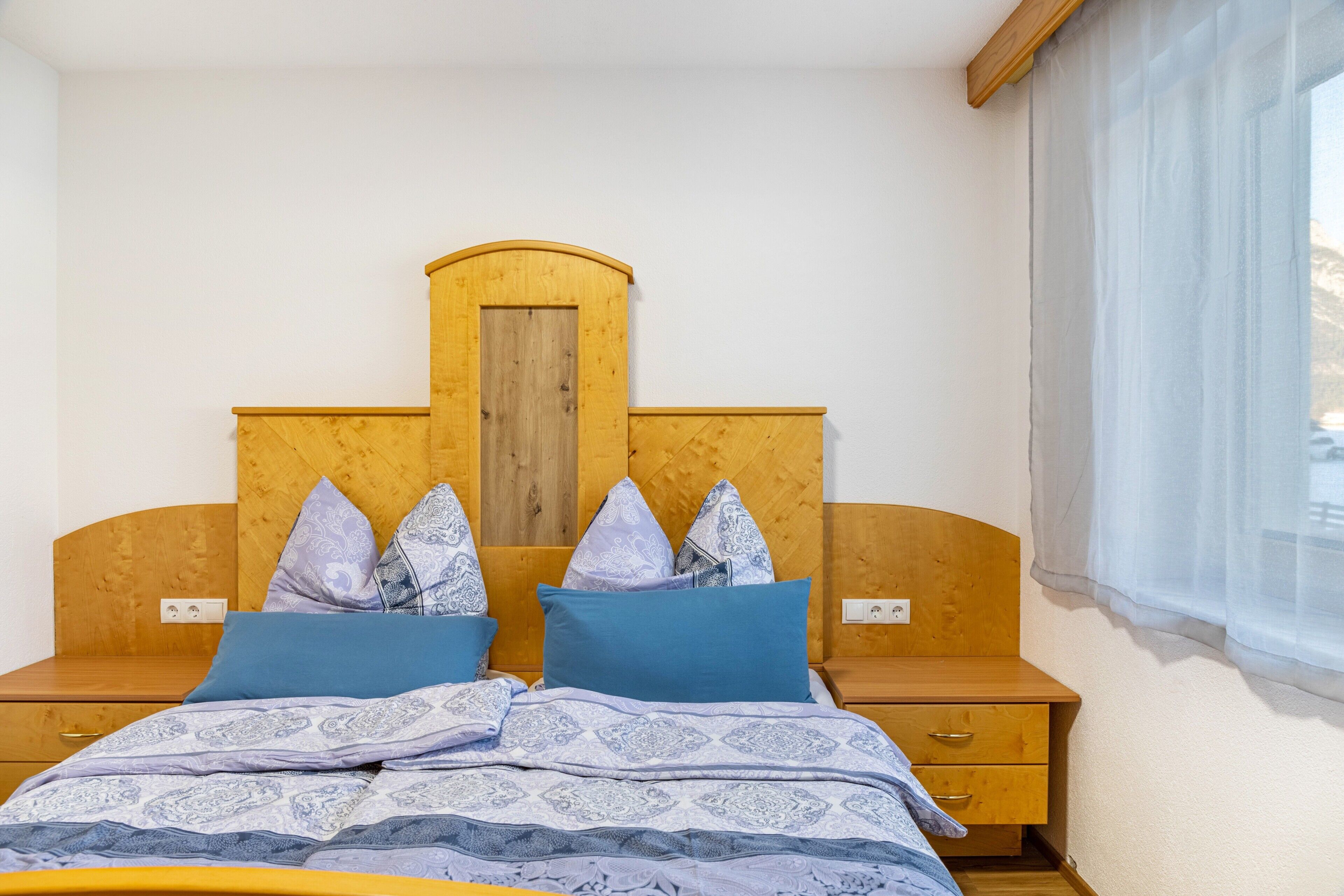 Una camera da letto con una testiera in legno, due comodini, un letto con biancheria a motivi e una finestra con tende trasparenti.