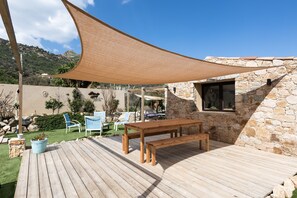 A wooden deck with a table and chairs, a shade sail, and a stone building with a window. - Room 'Lotu' with Mountain View, Wi-Fi and Air Conditioning (Santa-Reparata-di-Balagna)