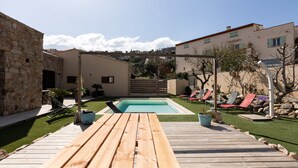 Un pontile in legno conduce a una piscina, circondata da edifici in pietra e da un'area verde con poltrone relax.