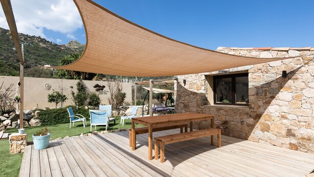 Un terrazzo in legno con tavolo e sedie, un telo ombreggiante e un edificio in pietra con una finestra.