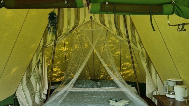 Una tienda de campaña con una cama, una silla verde, una mesa de madera con una cafetera y una alfombra estampada.