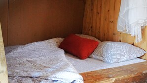 A small, wooden cabin-style room with a single bed, a red pillow, and a patterned pillow.