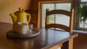 A wooden table with a yellow teapot, two cups, and a glass on a coaster. A wooden chair is behind the table, and a window with a view of greenery is visible.