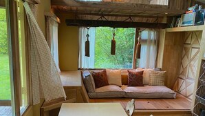 A cozy room with a wooden floor, a white table, a sofa with cushions, and a window with curtains. - Camping Ont Moeten - Hallertau (Rekken)