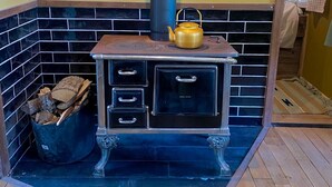 A wood-burning stove with a black chimney, a yellow kettle on top, and a black trash bin filled with wood logs. - Camping Ont Moeten - Hallertau (Rekken)