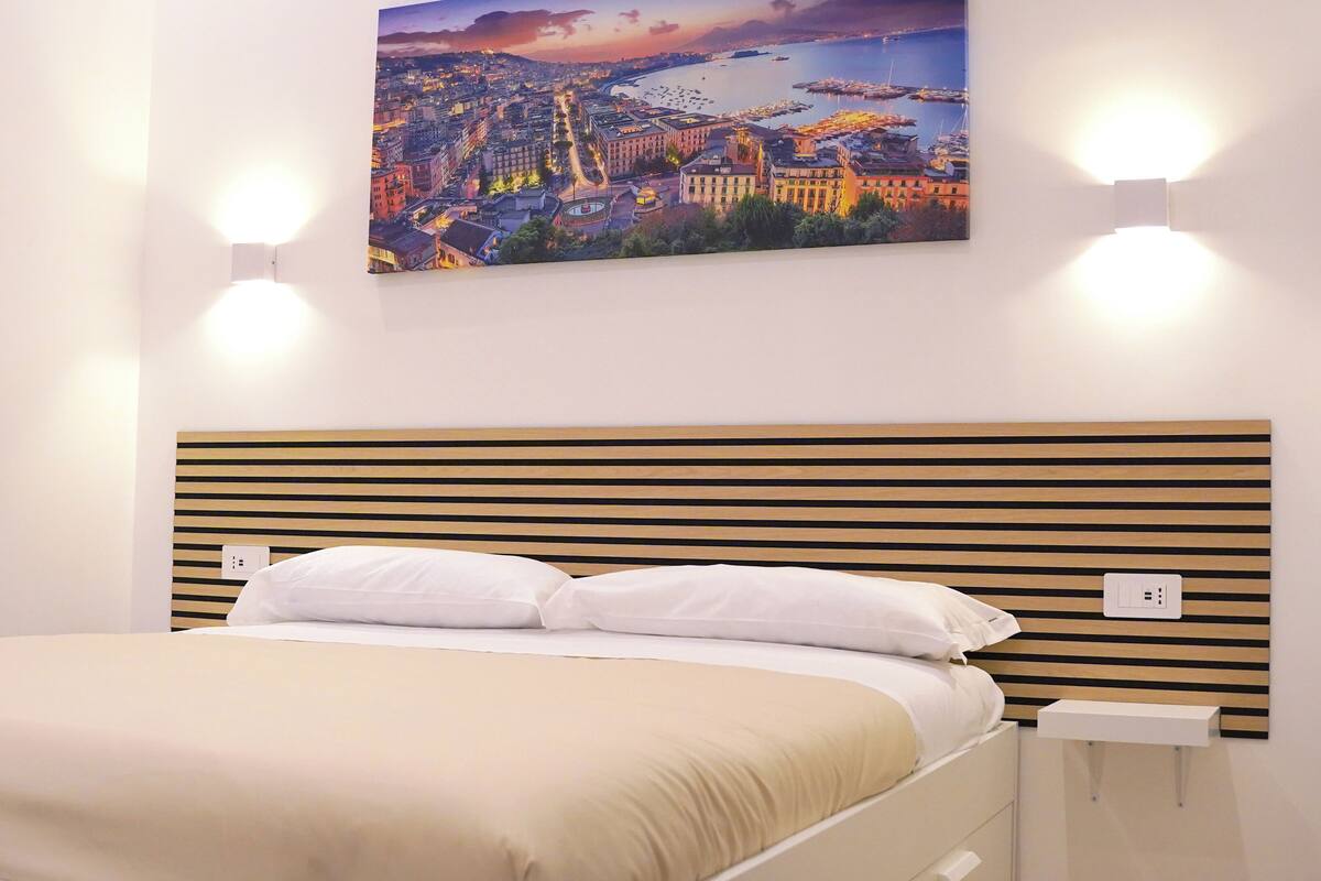 A neatly arranged hotel room with a bed, a headboard with horizontal wooden slats, and a cityscape painting above the bed.