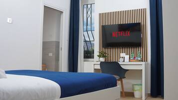 A hotel room with a bed, a desk with a computer, a TV displaying "NETFLIX", a window with a view of greenery, and a small trash bin.