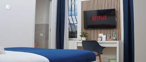 A hotel room with a bed, a desk with a computer, a TV displaying "NETFLIX", a window with a view of greenery, and a small trash bin.