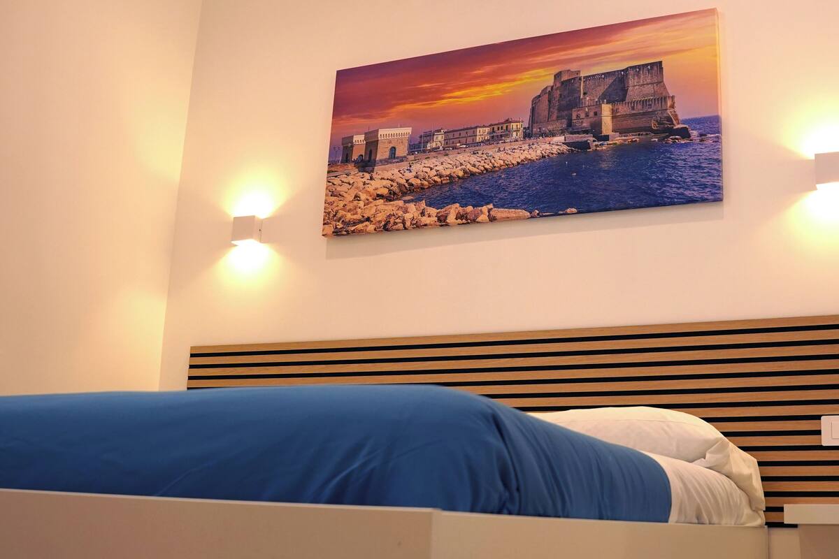 A bedroom with a bed, a headboard with horizontal slats, and a painting of a coastal scene above the bed.