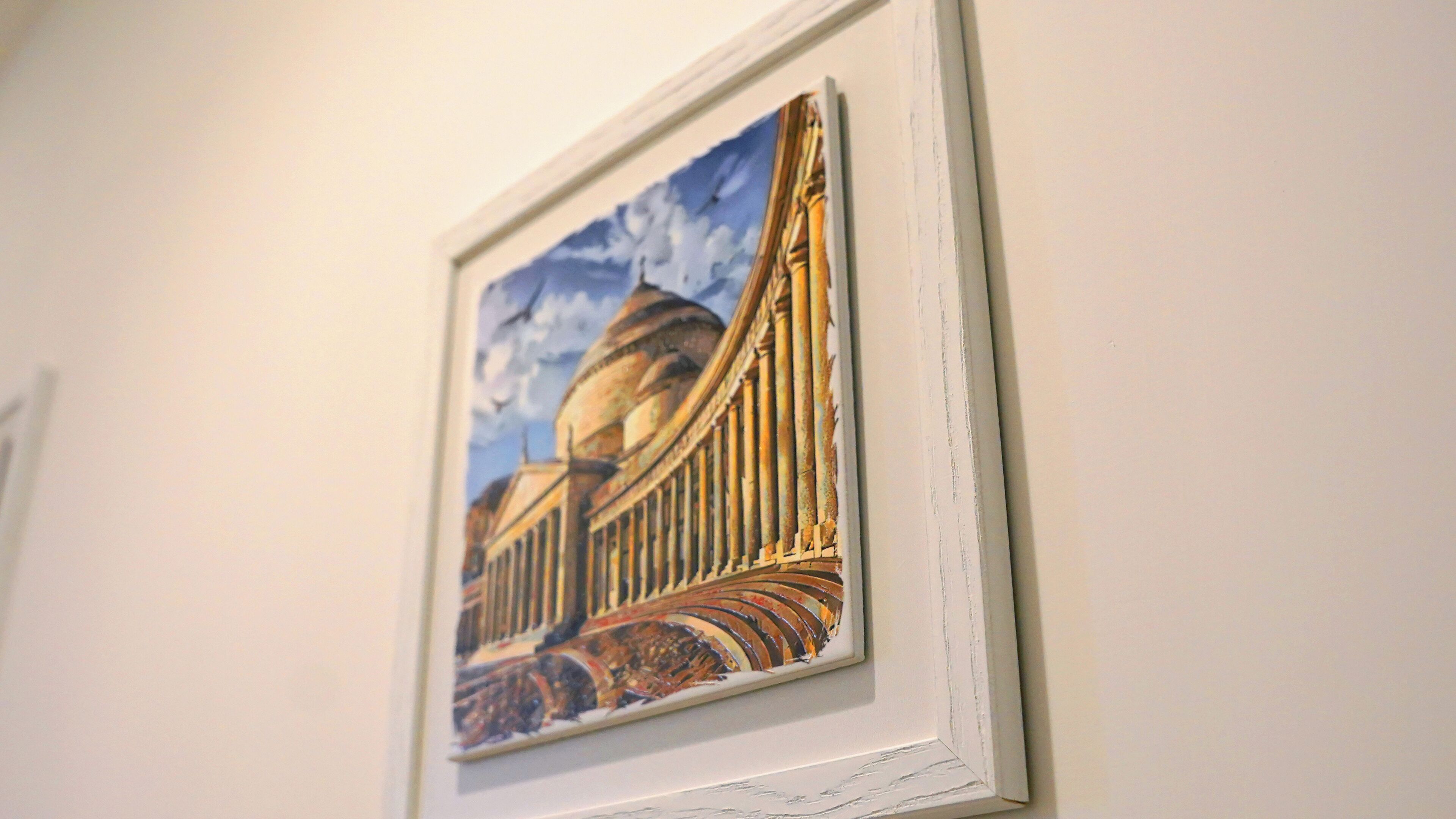 A framed artwork of a building with columns and a dome, mounted on a white wall.