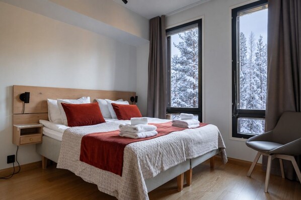 Une chambre moderne avec un lit, des tables de chevet, une chaise et de grandes fenêtres donnant sur un paysage enneigé.