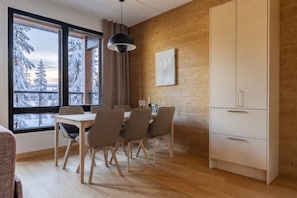 Un espace repas moderne avec une table et des chaises en bois, un luminaire suspendu noir et une vue sur des arbres enneigés à travers de grandes fenêtres.