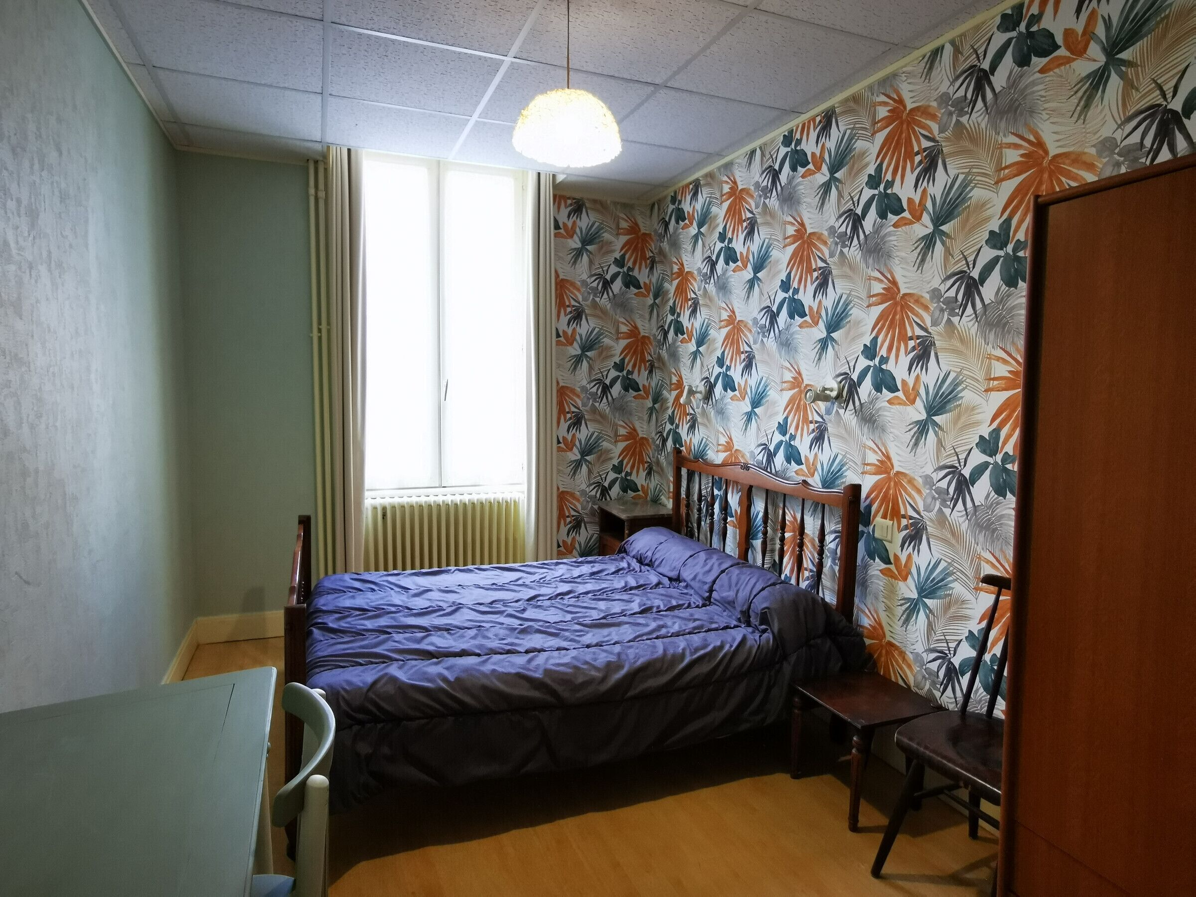 Un dormitorio con cama, una silla, un escritorio, un radiador y una ventana.