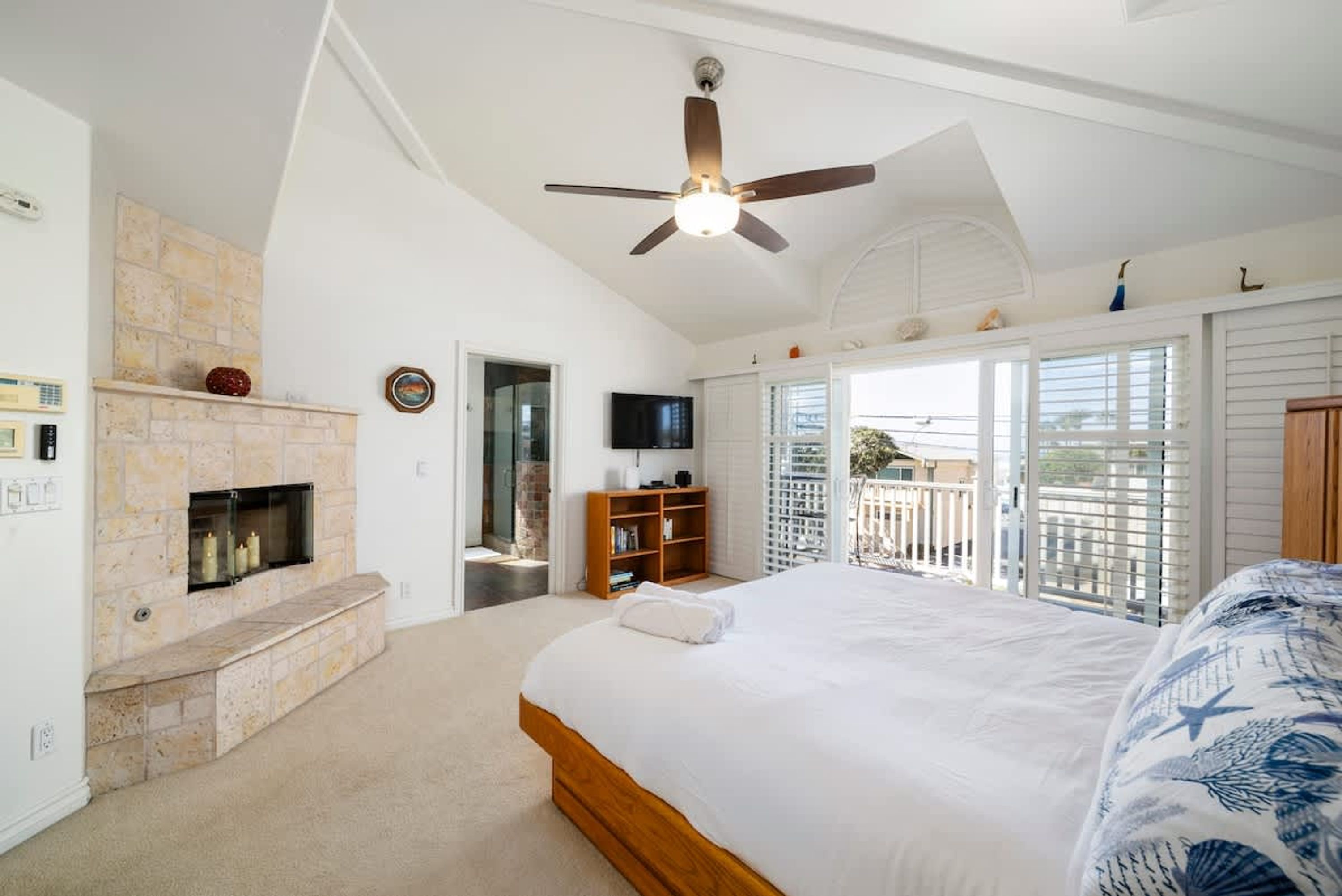 A bedroom with a large bed, a fireplace, a ceiling fan, and a balcony with a view.