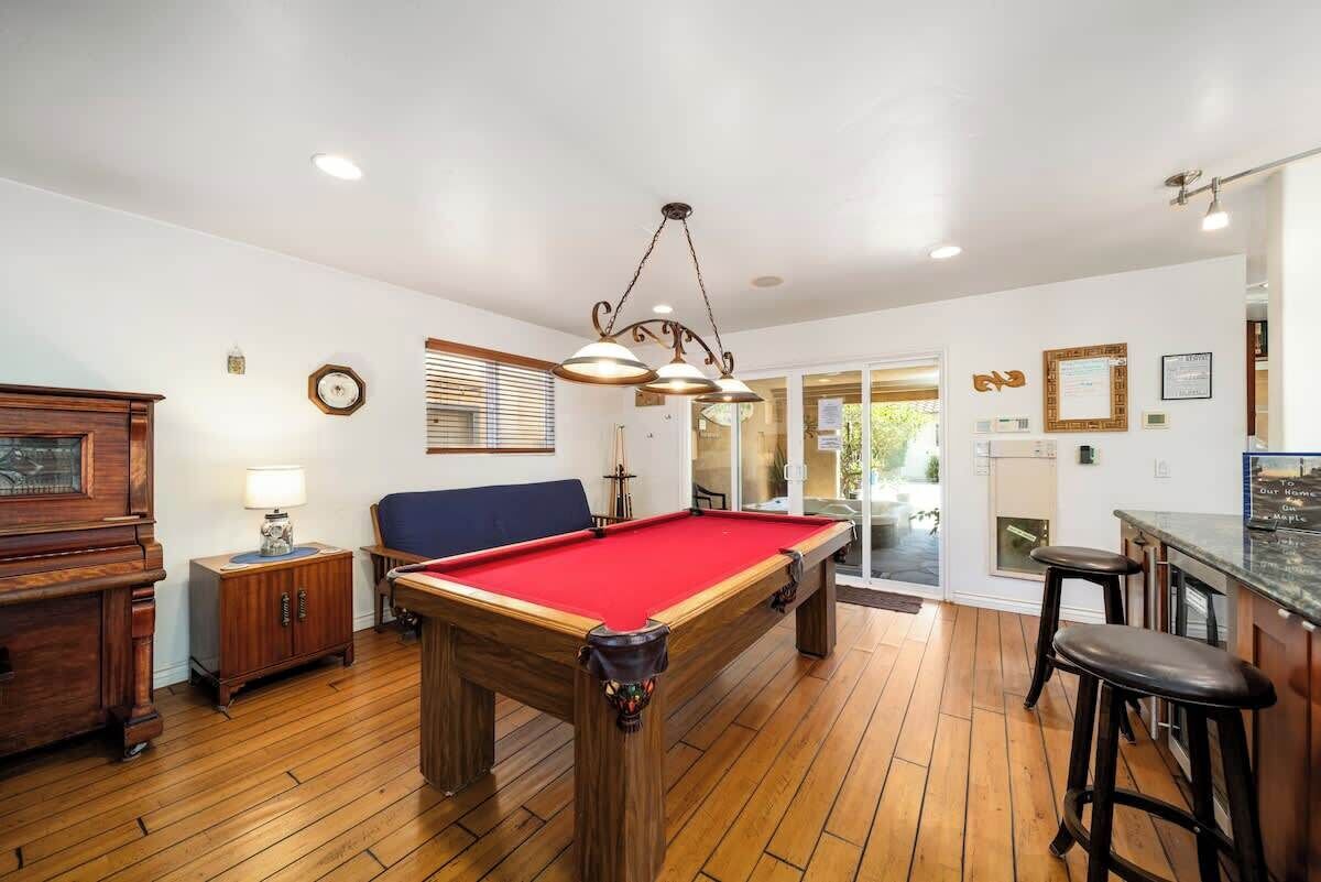 A pool table in a room with wooden flooring, a bar area with stools, and a ceiling light fixture.