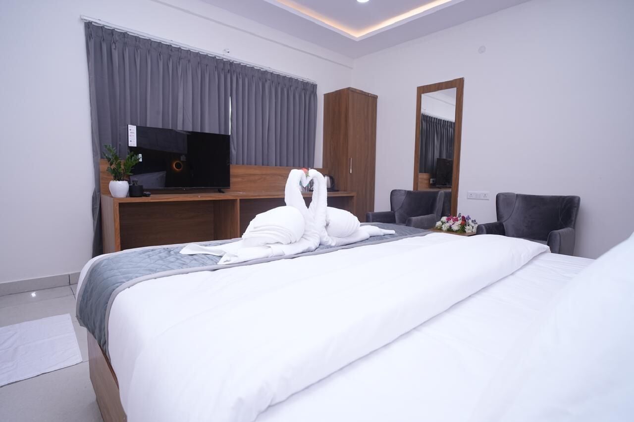 A hotel room with a neatly made bed, a television on a wooden stand, a mirror, and a grey chair.