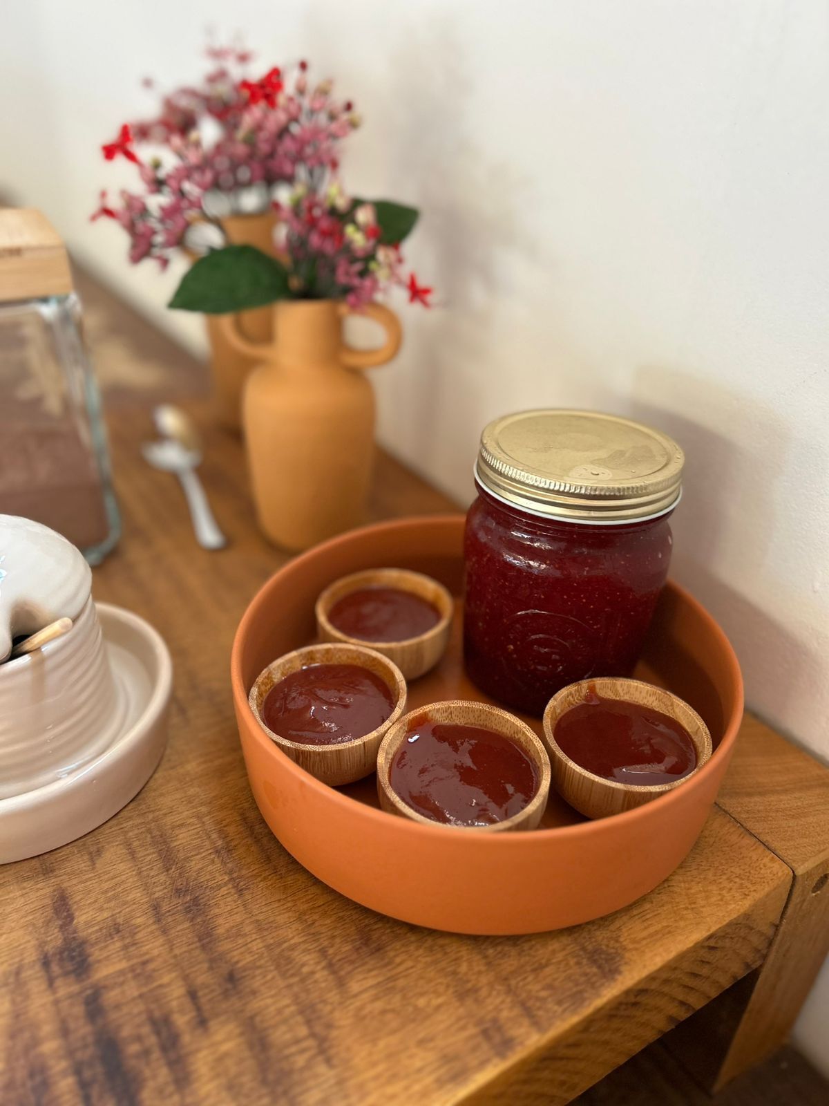 A wooden table with a terracotta bowl containing a jar of jam and small wooden cups.