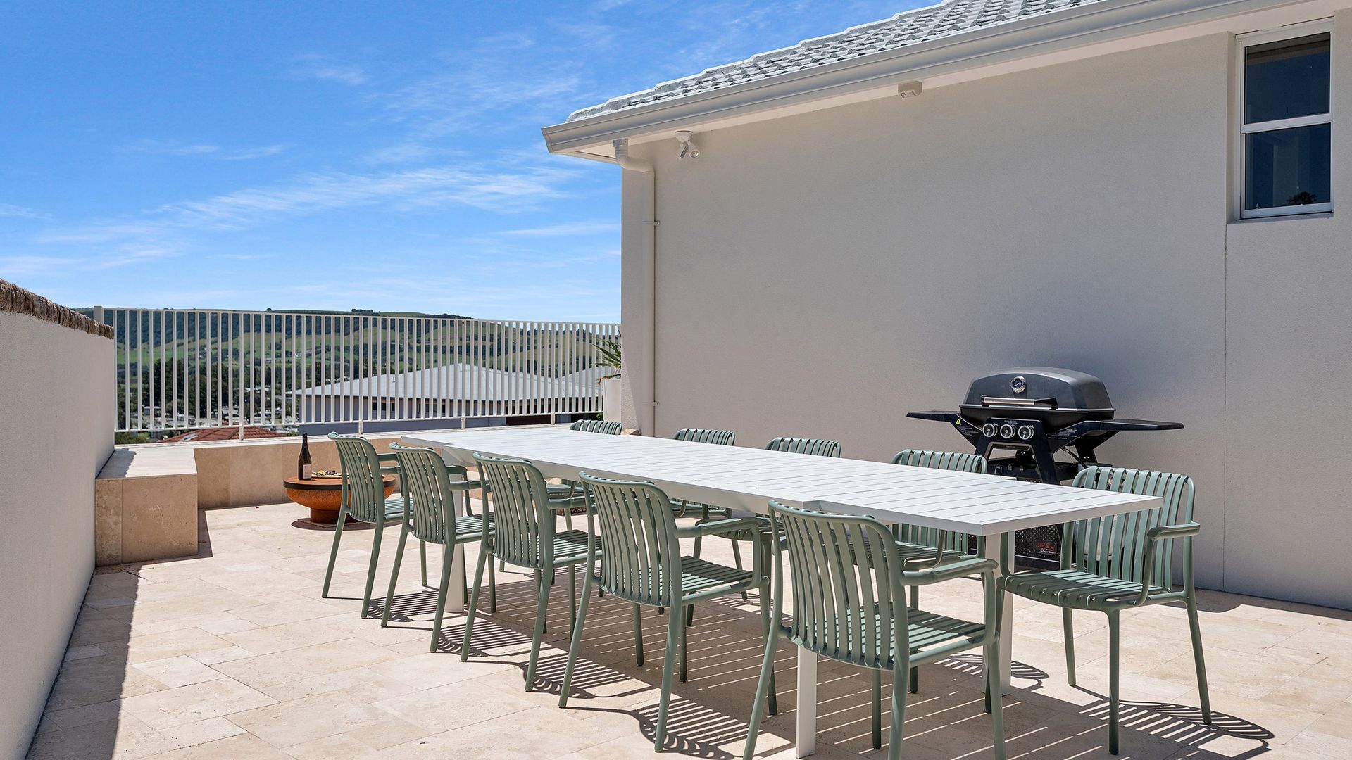 An outdoor patio with a long table and chairs, a grill, and a view of the hills.