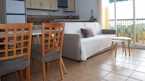 Une cuisine moderne avec des meubles bleus, un coin repas avec des chaises et une table en bois, un canapé gris et un balcon avec des chaises vertes.