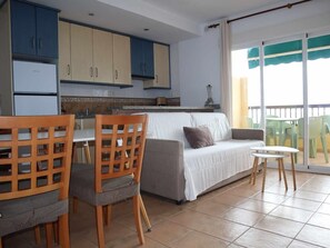 A modern kitchen with blue cabinets, a dining area with wooden chairs and a table, a grey sofa, and a balcony with green chairs. - Bright seaside apartment in Almuñécar (Almuñécar)