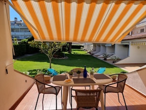 An outdoor patio with a striped awning, a round table, wicker chairs, and a view of a well-maintained lawn and buildings in the background. - Apartment with terrace 400m from Riells beach (L'Escala)