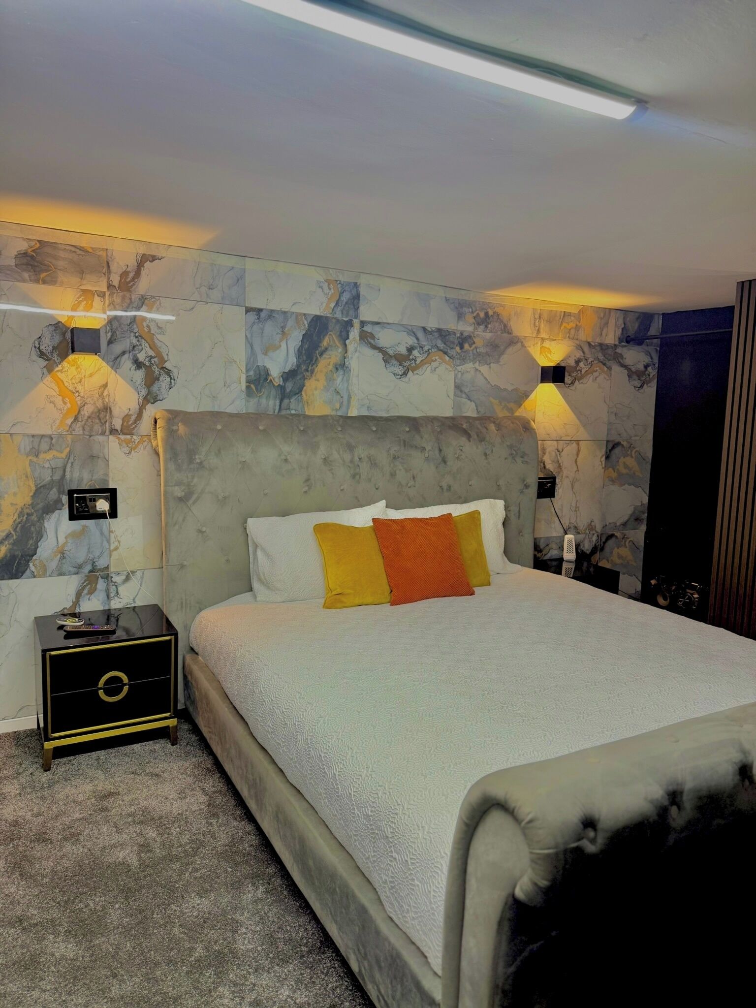 A modern bedroom with a marble-patterned wall, a large bed with white and orange pillows, and two wall-mounted lamps.