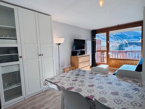 Ein Zimmer mit Esstisch, Stühlen, Fernseher, Schrank und einem Balkon mit Bergblick.