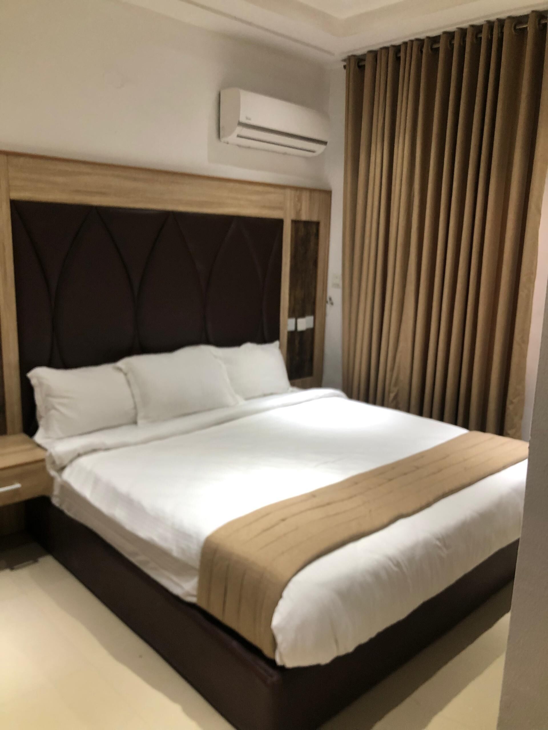 Une chambre d’hôtel avec un grand lit, une tête de lit en bois, une table de chevet, un appareil de climatisation et une fenêtre avec des rideaux.