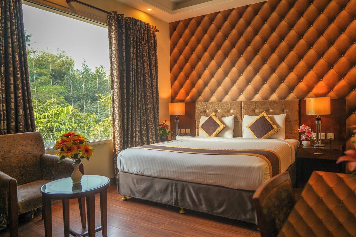 A hotel room with a large bed, a sitting area with a chair and a small table, and a view of greenery outside.