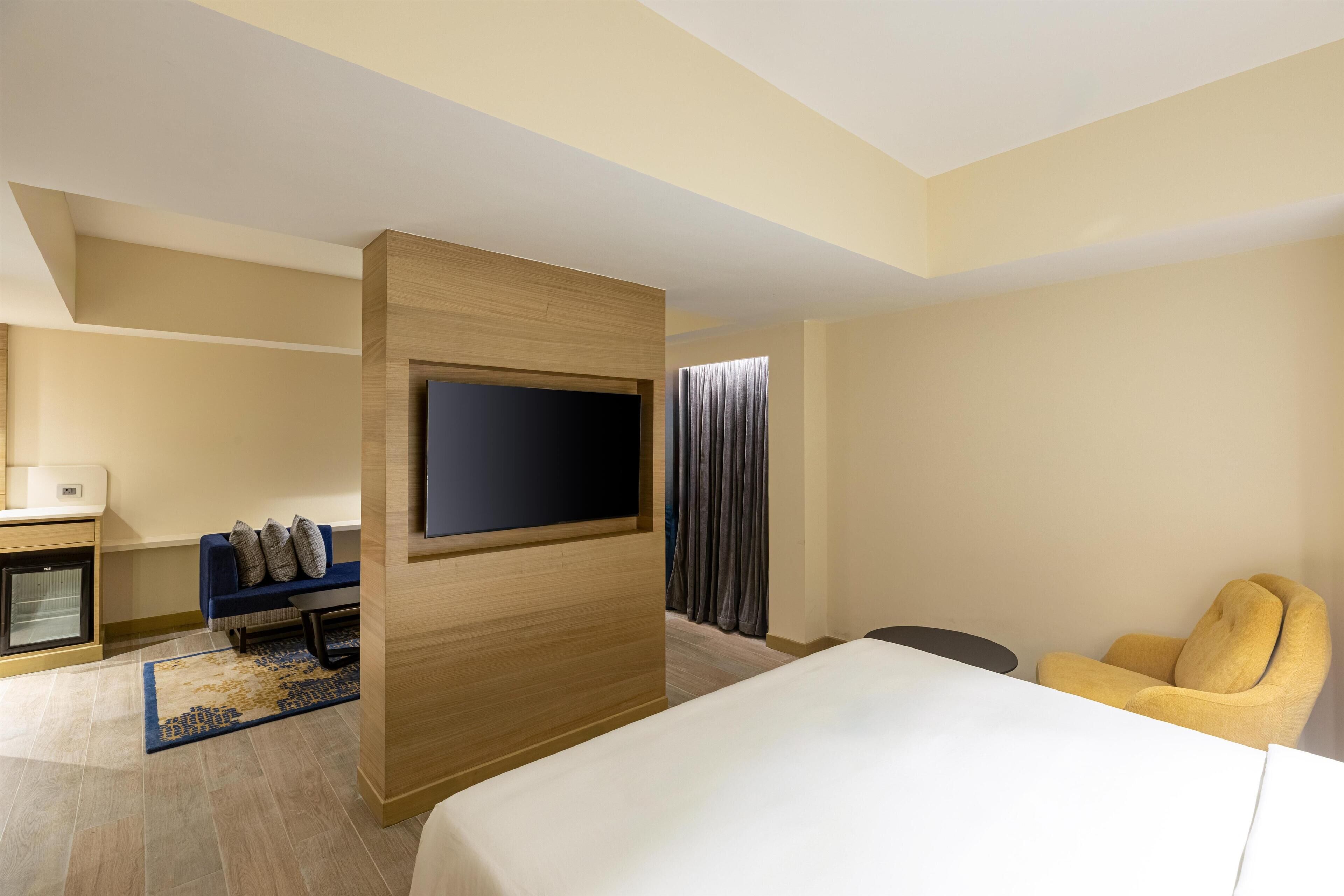 Et hotelværelse med en seng, et fjernsyn monteret på en træpanel, en sofa med puder, et lille bord og en stol.