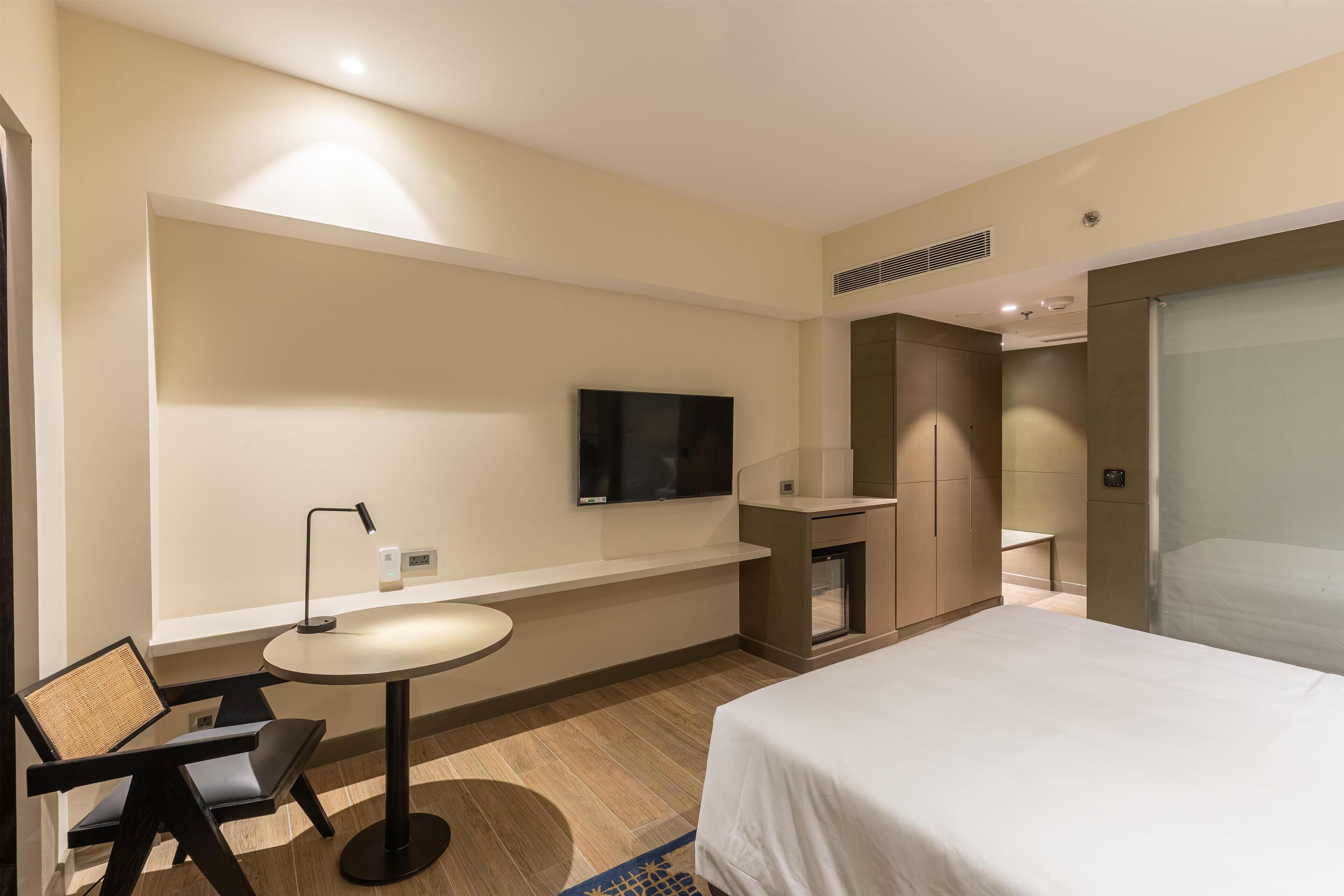 A hotel room with a bed, a television mounted on the wall, a small round table with a chair, and a built-in bench.