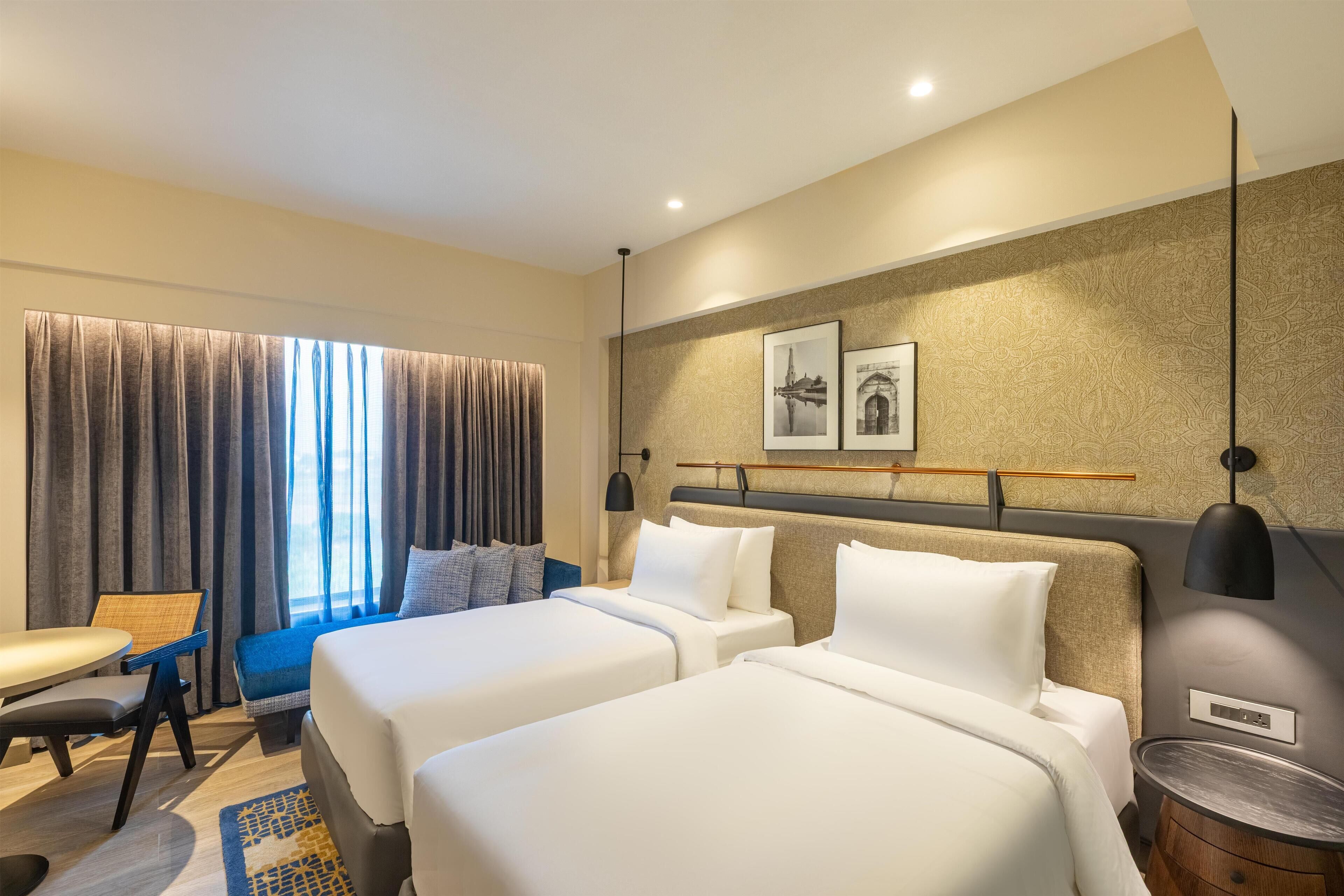 A hotel room with two beds, a desk, and a chair. There is a window with curtains, a wall-mounted lamp, and a framed picture.