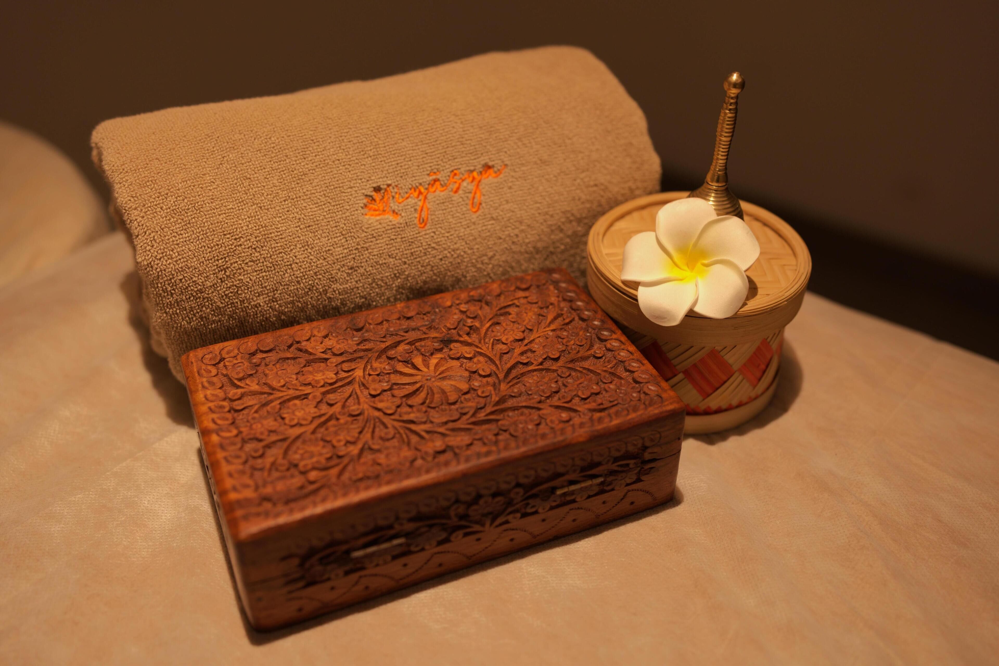 A brown textured box with floral carvings, a brown towel with orange embroidery, and a wooden bowl with a white flower and a spoon.