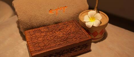 A brown textured box with floral carvings, a brown towel with orange embroidery, and a wooden bowl with a white flower and a spoon.
