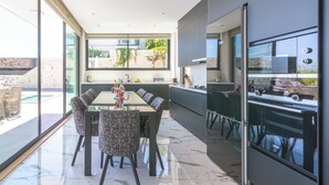 A modern kitchen with a large island, a dining area with a glass table, and a view of a swimming pool. - Eco Villa Mirada (Kalkan)